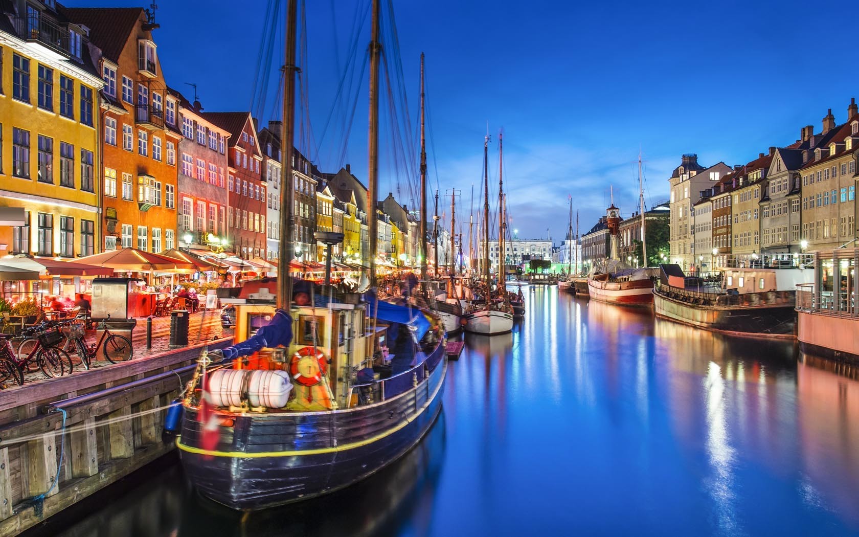 Colorful waterfront buildings along a canal at dusk, with boats moored in the water and city lights reflecting on the surface, evoking Copenhagen's atmosphere